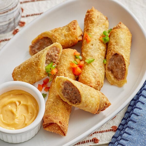 Crispy FATBOYSBBQ egg rolls filled with meat, garnished with peppers, served with a side of dipping sauce.