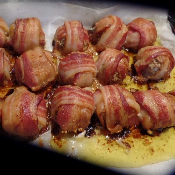 Close-up of bacon-wrapped meatballs or scallops, glistening with rendered fat, on a baking sheet.