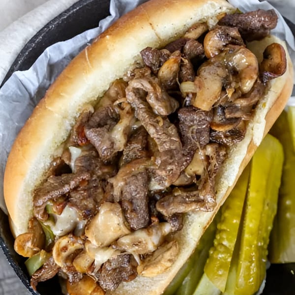 A FATBOYSBBQ cheesesteak sandwich with steak, mushrooms, and melted cheese, served with pickle spears.