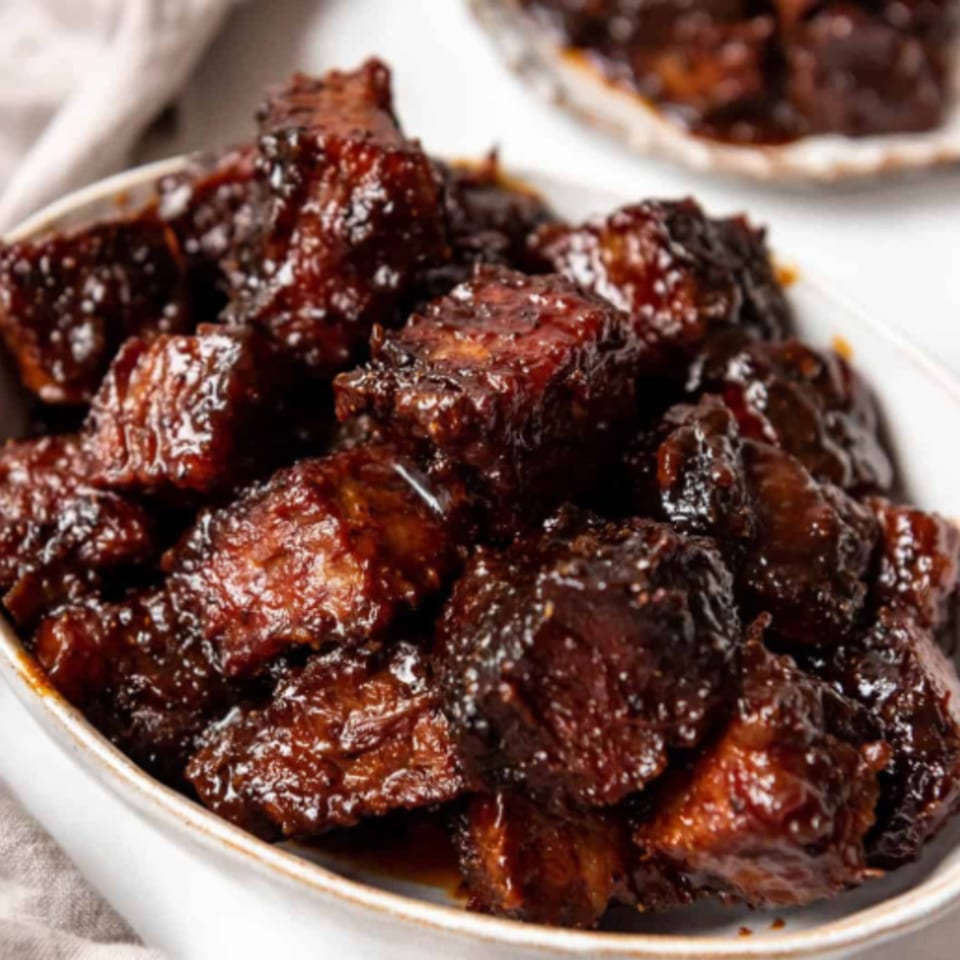 Close-up of FATBOYSBBQ burnt ends, glistening with dark, rich barbecue sauce in a white bowl.